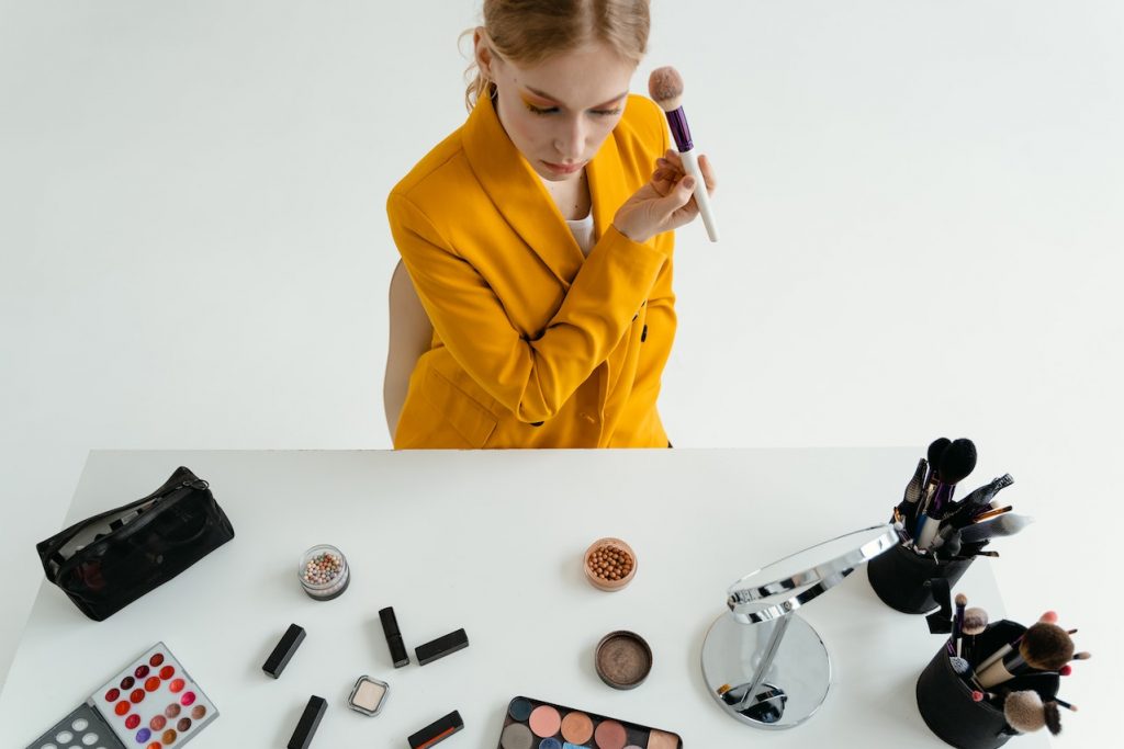 Woman wore a orange color top doing makeup having all cosmetics on white table
